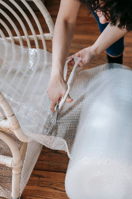 Interior scene showing a home relocation process with several cardboard moving boxes stacked on a wooden floor near a doorway. A few boxes are wrapped in clear plastic and secured with packing tape, i