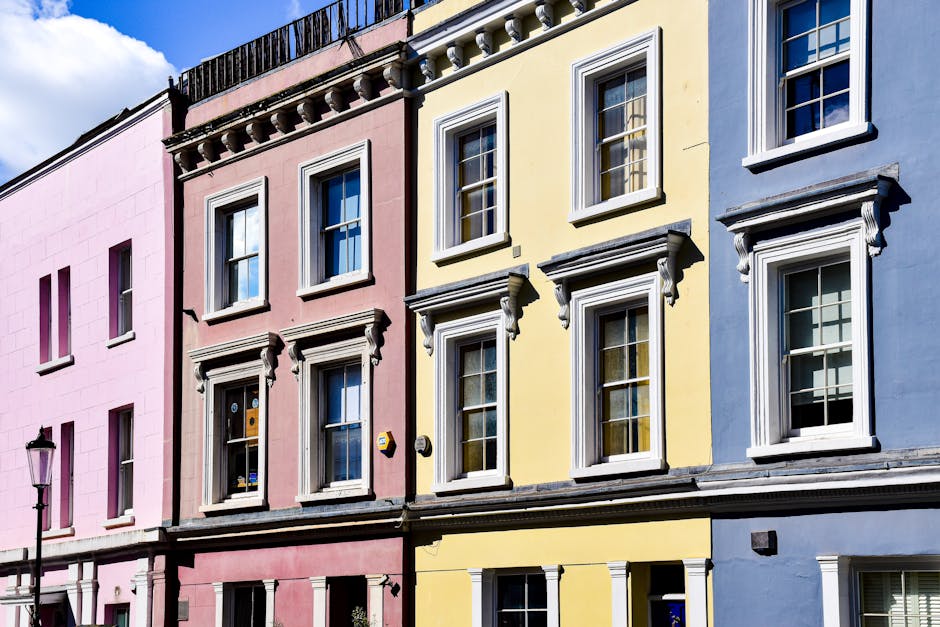 A row of multi-storey terraced houses on a residential street in Notting Hill, London, with each building featuring distinct exterior colours and architectural details. The buildings display a mix of 