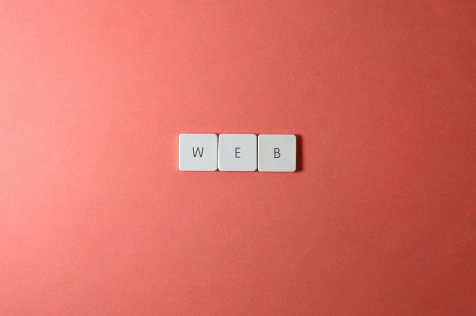 Close-up image of three white square tiles with black letters spelling 'WEB' arranged horizontally on a smooth, pastel pink background. The tiles are positioned near the center, with even spacing betw